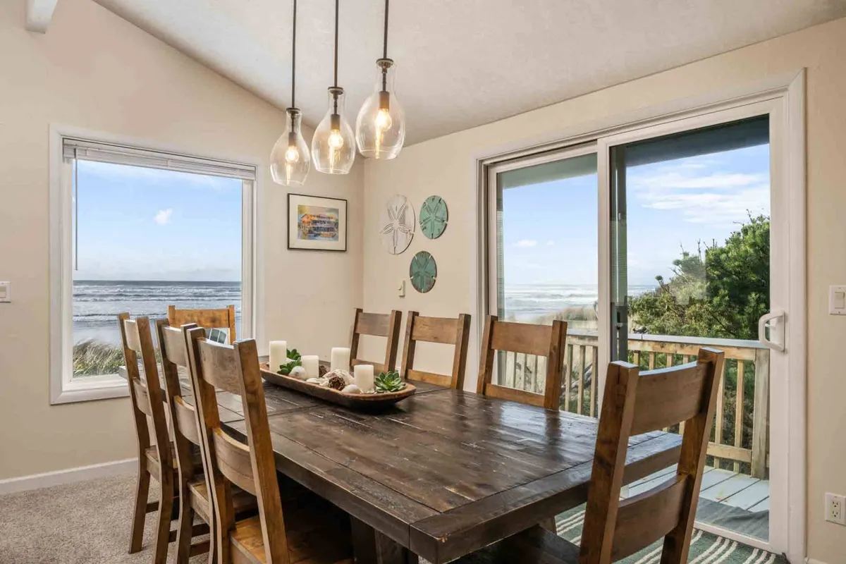 A spacious dining area featuring a large wooden table set beneath stylish hanging light fixtures. Floor-to-ceiling windows provide panoramic ocean views, bathing the space in natural light, while the table is set for a meal, inviting guests to gather and celebrate.