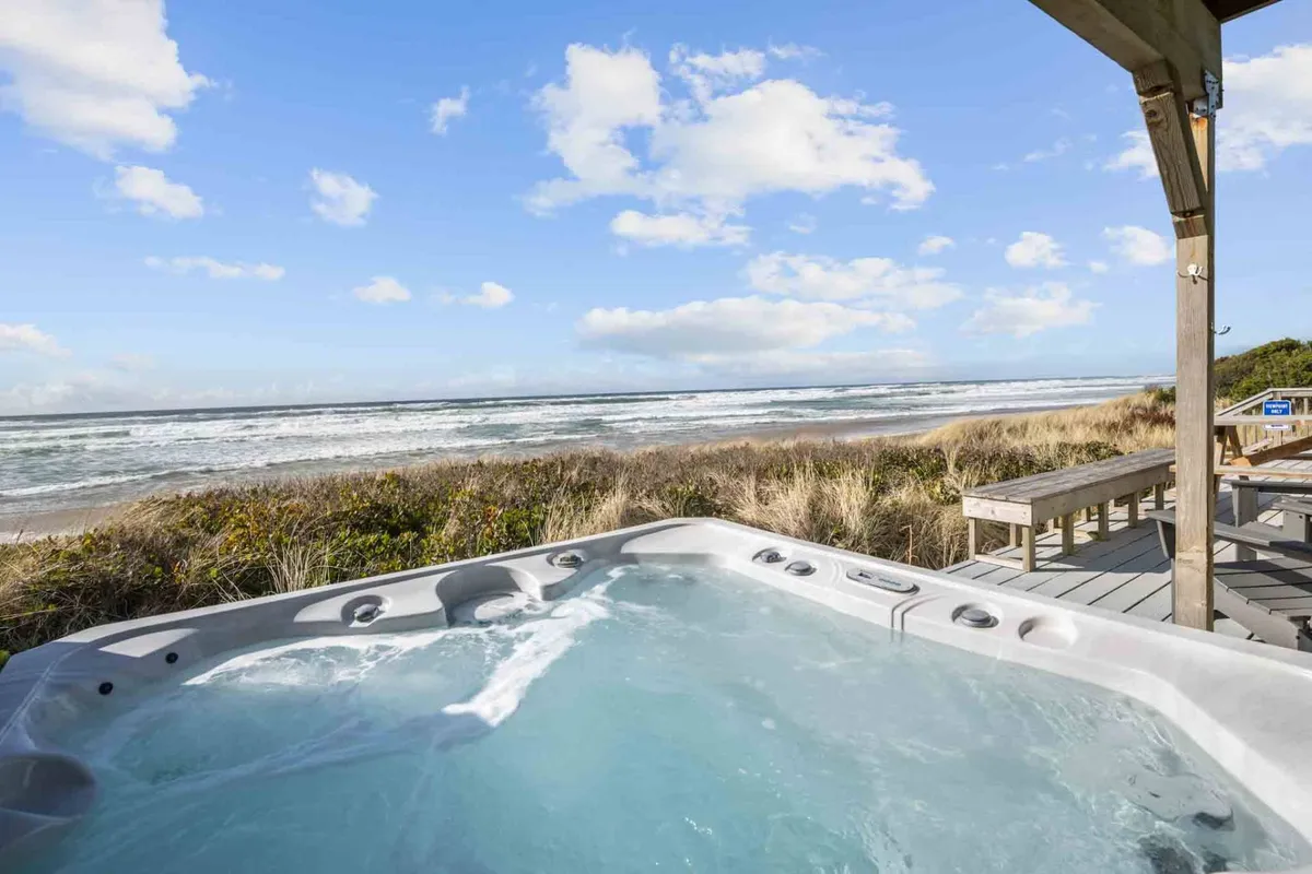 An inviting wooden deck overlooking the ocean, featuring a bubbling hot tub and open sky. The scene suggests relaxation and fun, with potential for socializing, soaking after a day of adventure, and unwinding as the sun sets.
