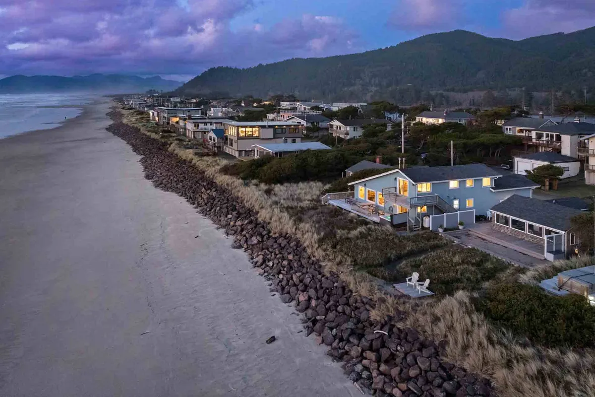 A panoramic exterior view of beachfront houses lining a rocky shoreline with gentle ocean waves and a stunning mountain backdrop. The image captures families walking along the beach, creating sandcastles, and gathering to enjoy the sunset. The scene is peaceful and vibrant, conveying the joy of seaside activities and natural beauty.