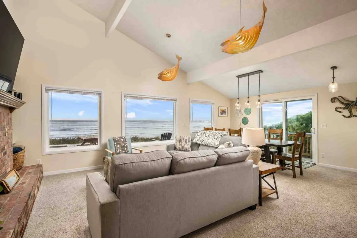 The image showcases a spacious, inviting living room with expansive windows framing stunning ocean views. A plush, comfortable sofa sits facing the windows, perfect for relaxing while watching the waves. Unique modern light fixtures add character, and the overall atmosphere is warm and welcoming, ideal for gathering with family and friends.