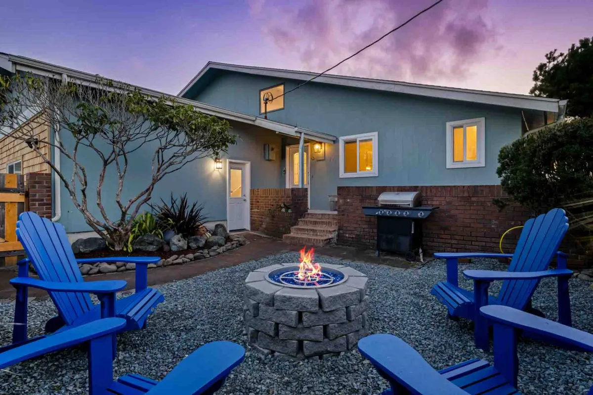 The exterior of the house with blue adirondack chairs gathered around a glowing fire pit, a grill nearby, and welcoming landscaping. The scene suggests communal evening gatherings, storytelling, and sharing meals outdoors under the stars.