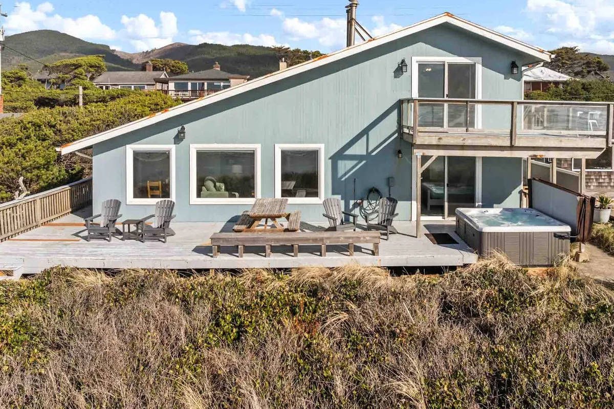 An inviting exterior showcasing the blue house, a spacious deck with seating, a hot tub, and lush surrounding greenery. The setting feels peaceful and private, perfect for outdoor relaxation and connection with nature.