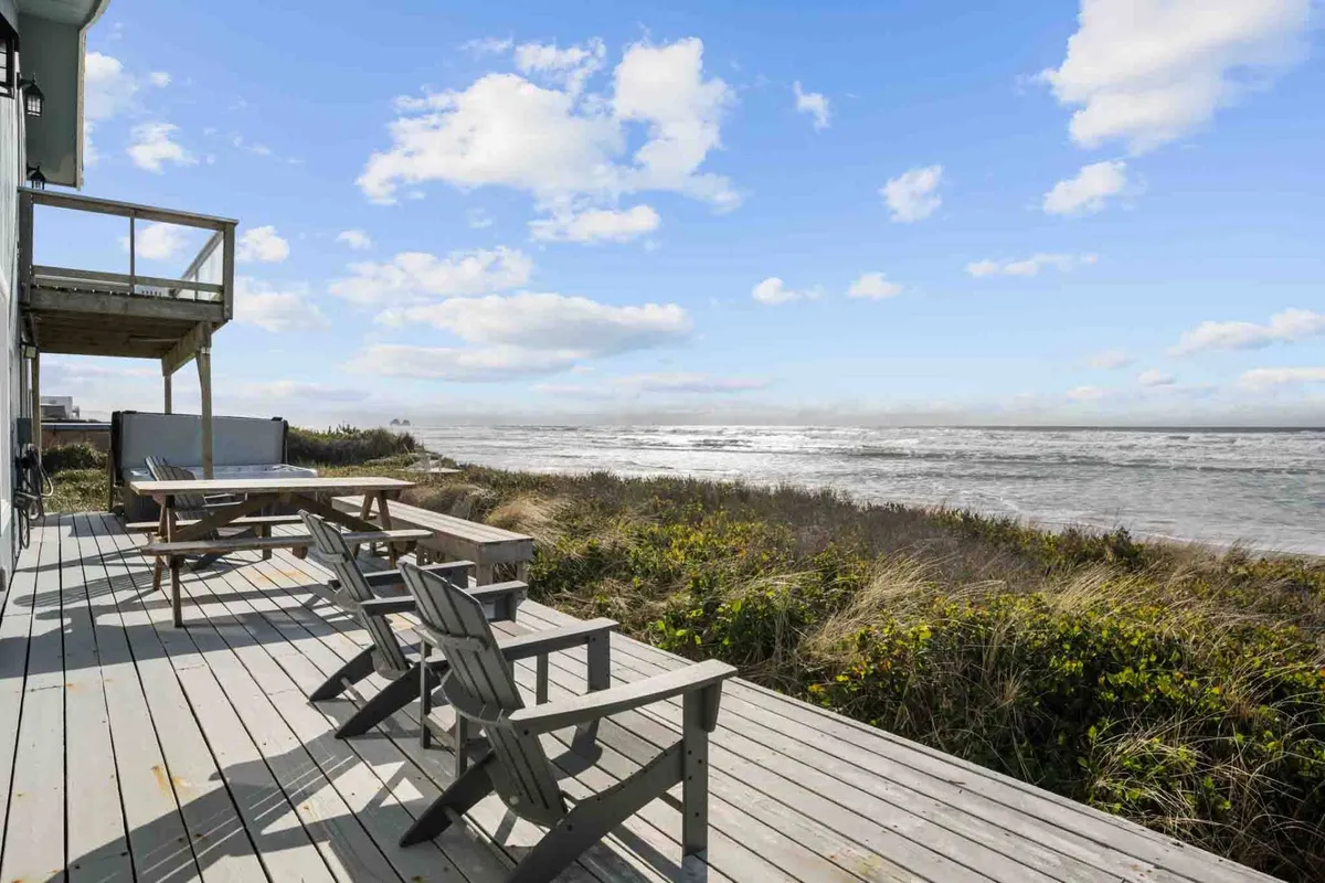 A spacious wooden deck overlooking the ocean, complete with comfortable chairs and a hot tub. Lush greenery surrounds the deck, enhancing the tranquil atmosphere. The hot tub is positioned for direct ocean views, and the seating area invites guests to relax and soak up sunsets or stargaze at night.