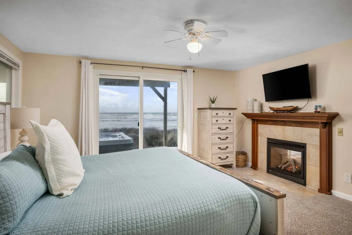 A cozy bedroom with a light blue quilted bed, sliding glass door opening to an outdoor view, a fireplace with decorative accents, and a wall-mounted TV. The atmosphere is serene, with an emphasis on comfort and indoor-outdoor living.