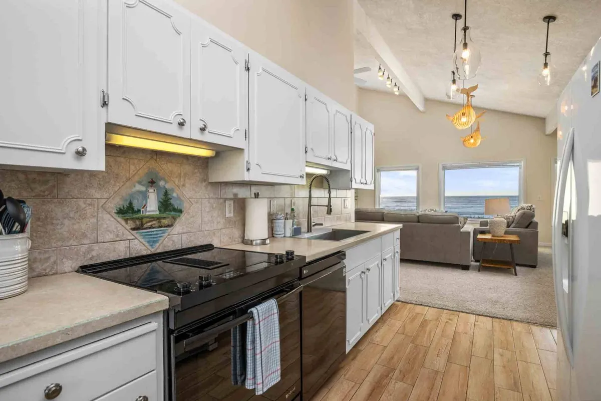 A bright, inviting kitchen with modern appliances, large windows letting in abundant natural light, a comfortable sofa nearby, and lighthouse-themed artwork above the counters. The kitchen overlooks the ocean, and the layout encourages both cooking and socializing.